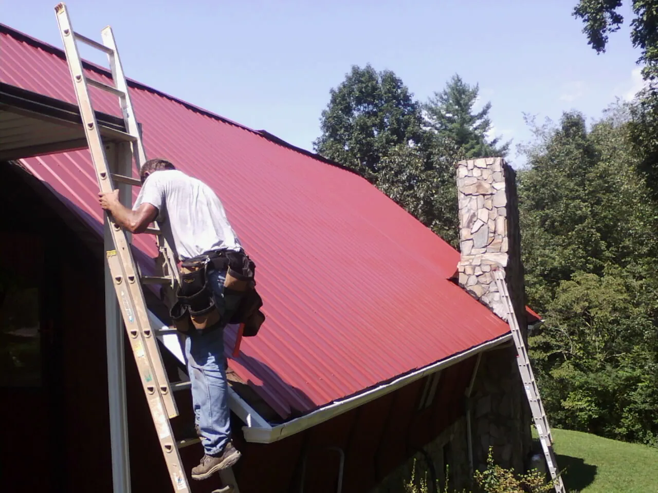 Roofer on a ladder beside a red metal roof and stone chimney — Soffit & Fascia Repair by Roofing Brothers in Wellesley Hills