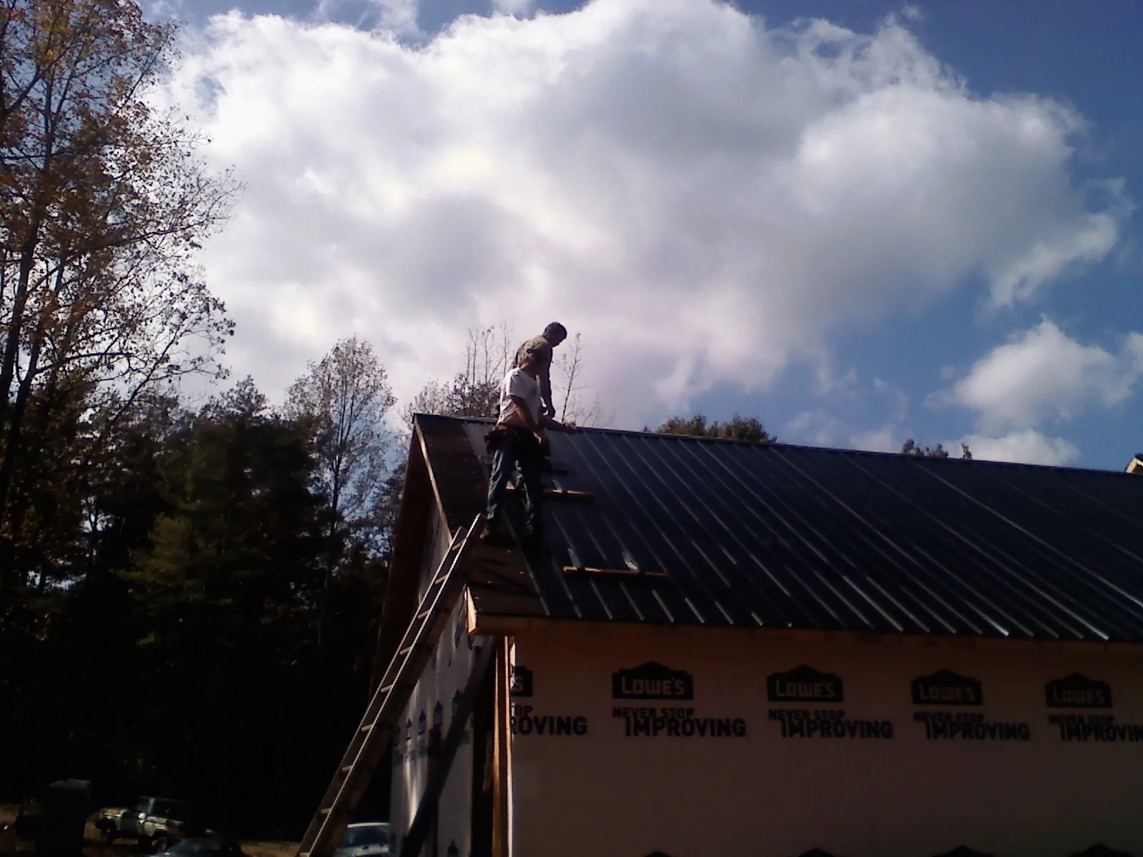 Roofer fastening dark metal panels high against a cloudy sky, from a Roof Inspection job in Wellesley Hills